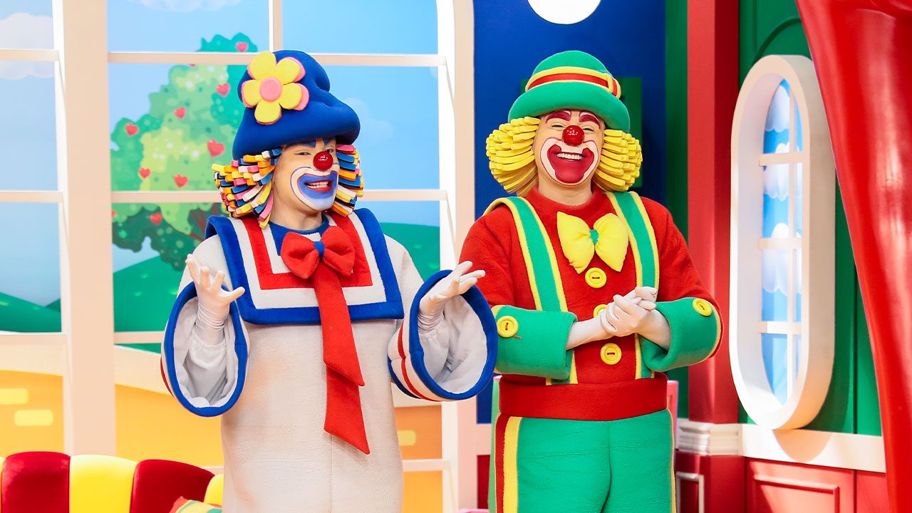 Patati e Patatá sorrindo no cenário colorido do Bom Dia & Cia com janelas e árvores ao fundo