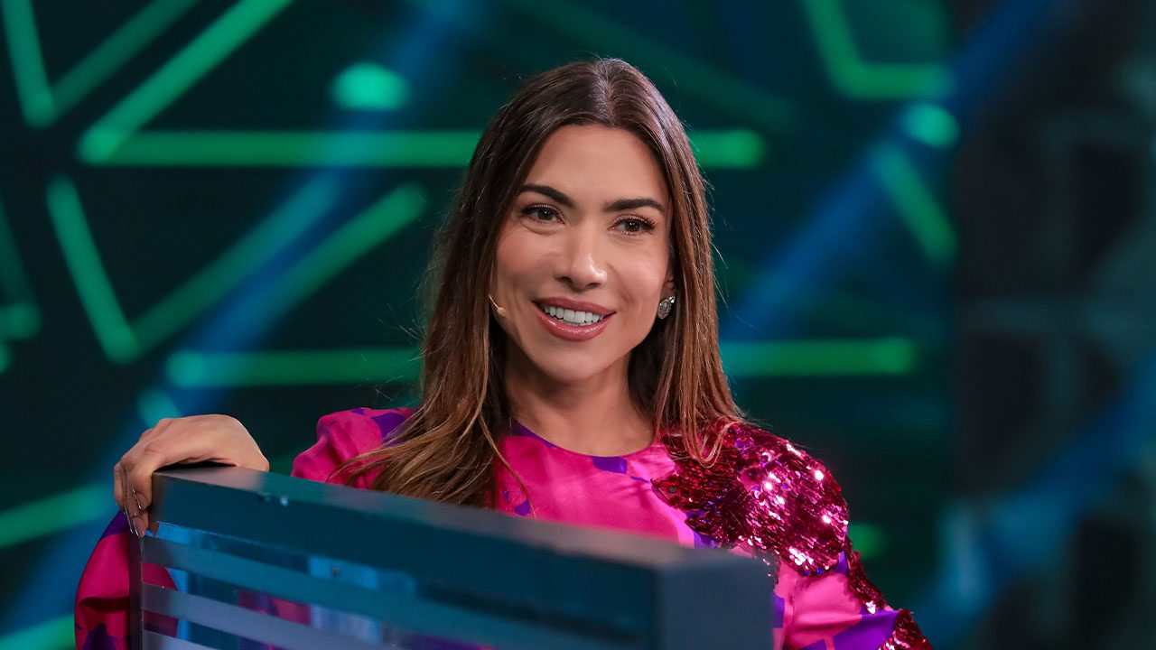 Patricia Abravanel sorrindo no palco do Programa Silvio Santos com figurino rosa e microfone no rosto