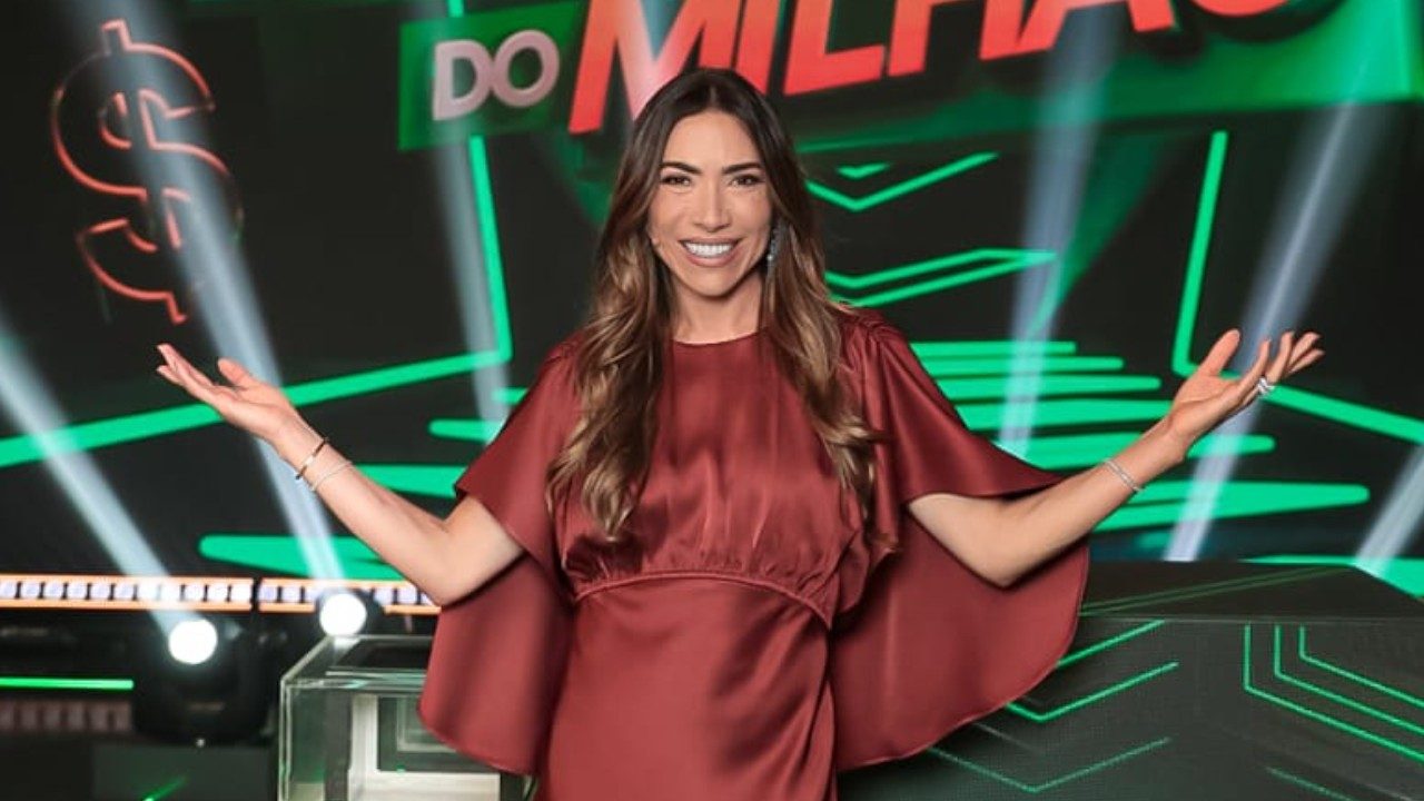 Patricia Abravanel abre os braços no palco do Show do Milhão com cenário verde e marca do programa ao fundo
