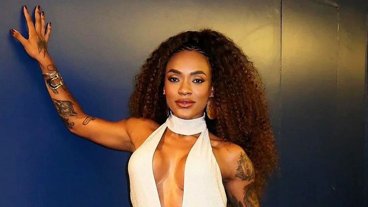 Patrícia Ramos posa com vestido branco de decote profundo e cabelos cacheados diante de parede azul escura