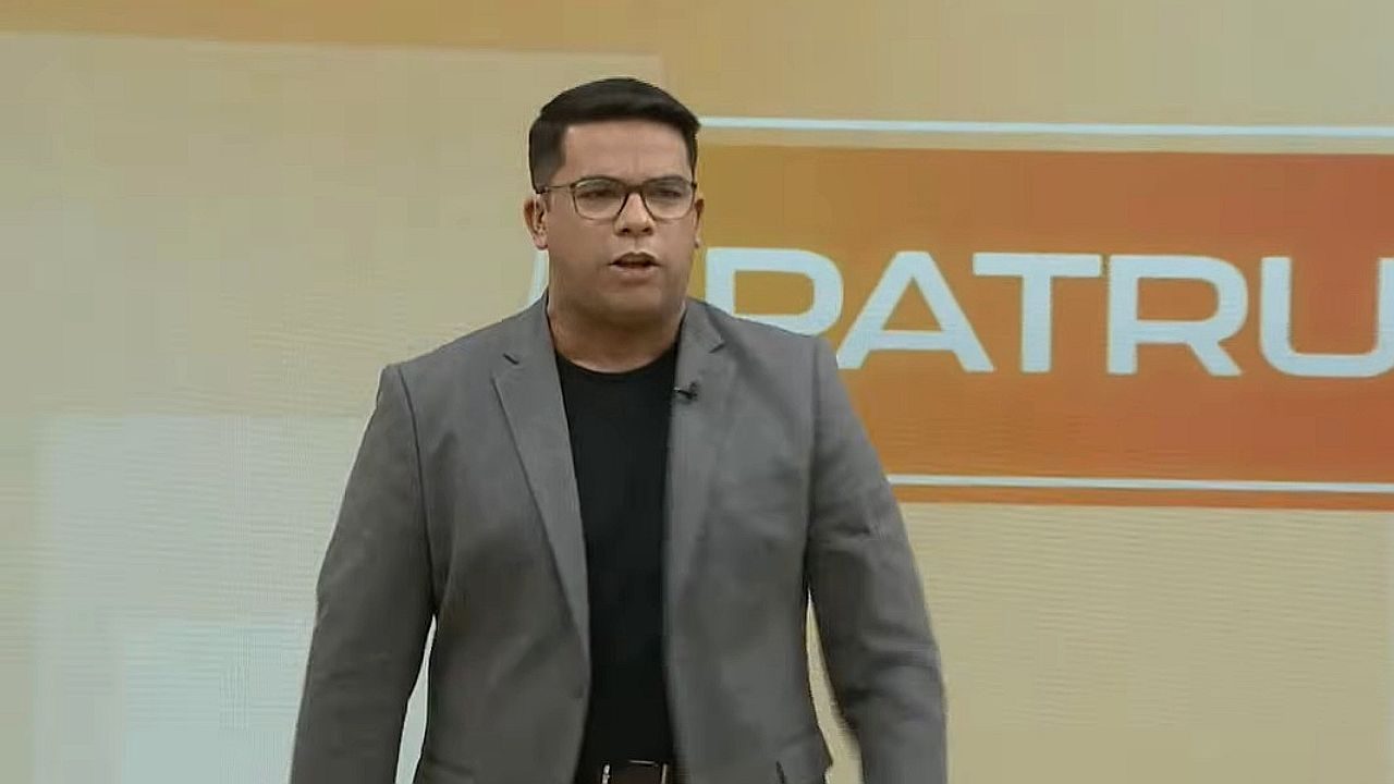 Apresentador de óculos e blazer cinza fala em estúdio de telejornal com painel laranja ao fundo exibindo programa Balanço Geral