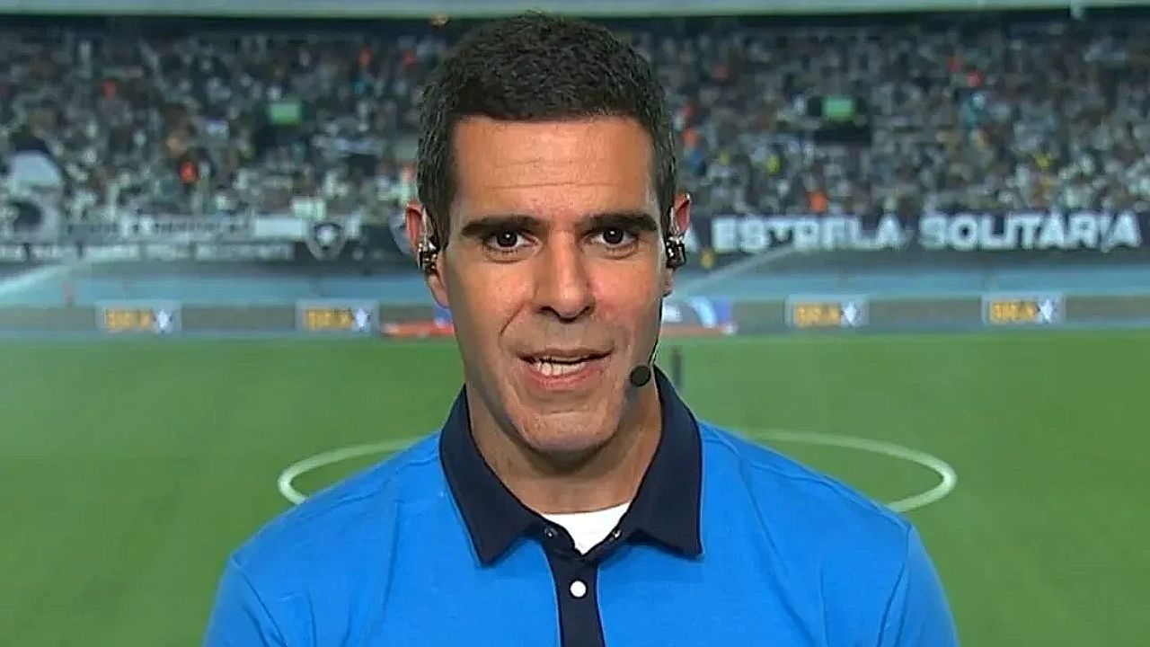 Repórter esportivo com microfone e ponto eletrônico fala diante de campo de futebol com arquibancada ao fundo