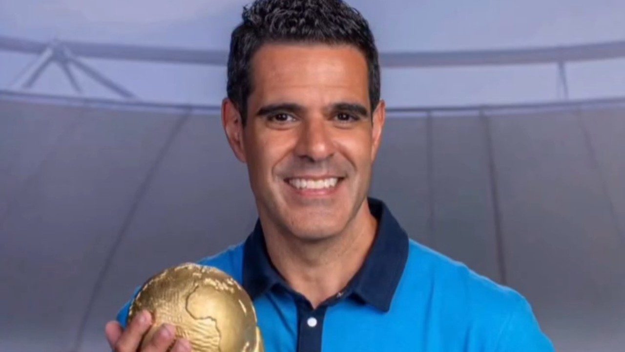 Paulo Andrade sorri segurando troféu em formato de globo usando camisa polo azul diante de cenário de estúdio esportivo