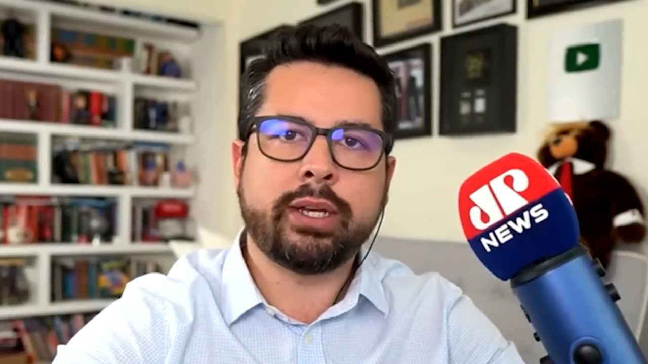 A imagem mostra Paulo Figueiredo, um homem usando óculos e camisa social clara, falando em frente a um microfone com o logotipo da Jovem Pan News. Ao fundo, há uma estante com livros e quadros decorativos, sugerindo um ambiente de home office