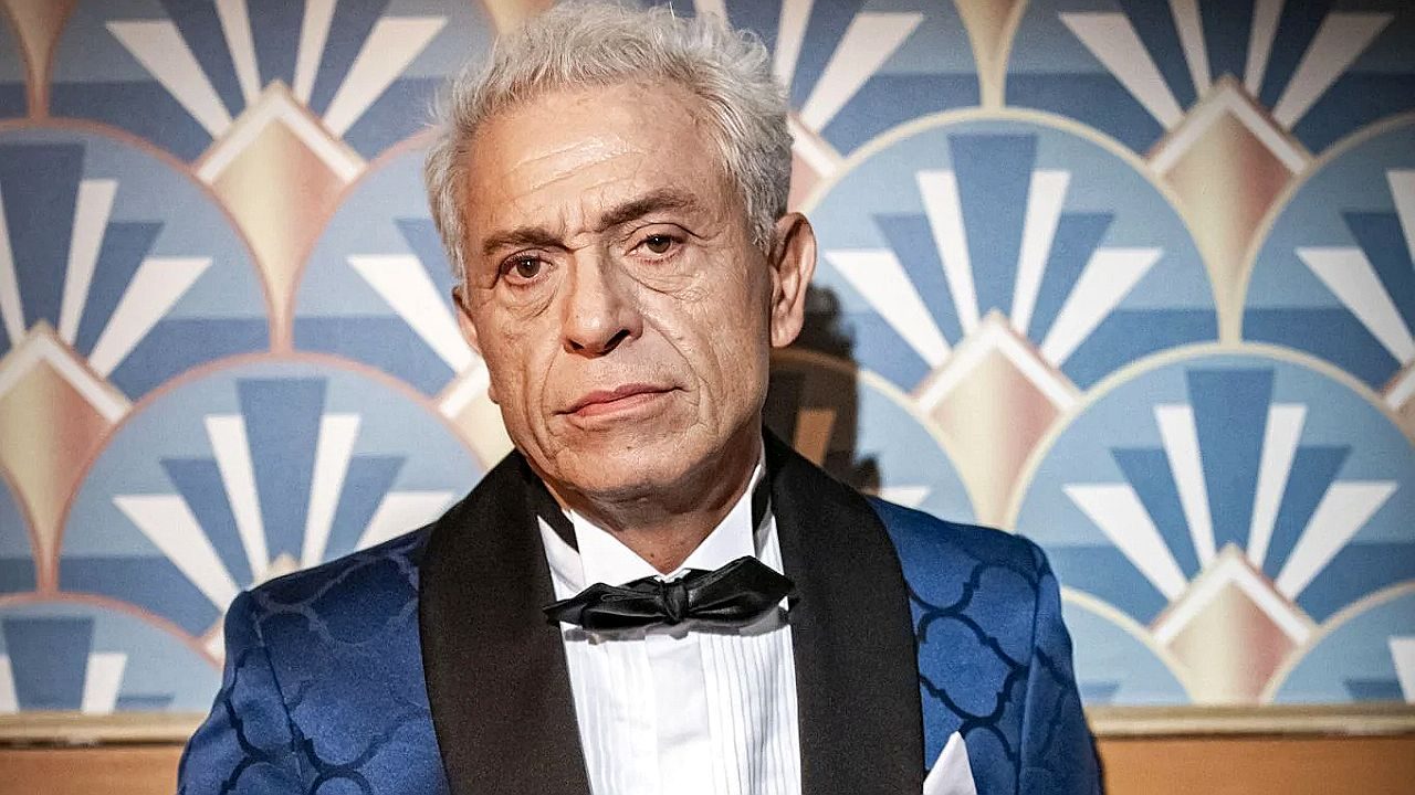 Homem de terno azul e gravata borboleta encara sério em cenário com decoração geométrica ao fundo