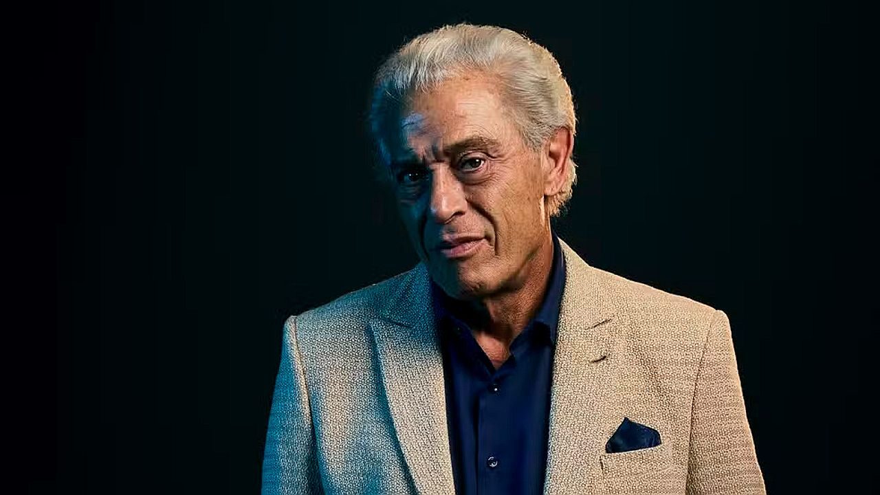 Paulo Gorgulho com blazer claro e camisa azul em retrato promocional com fundo escuro e iluminação dramática