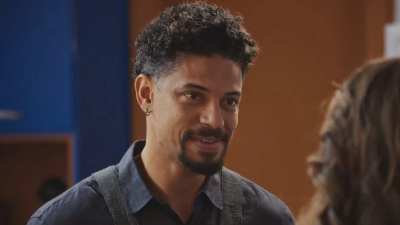 Homem jovem com barba e cabelo cacheado curto sorri levemente enquanto olha para outra pessoa fora de quadro; ele usa camisa azul e alças de suspensório em um ambiente interno com fundo azul e laranja.