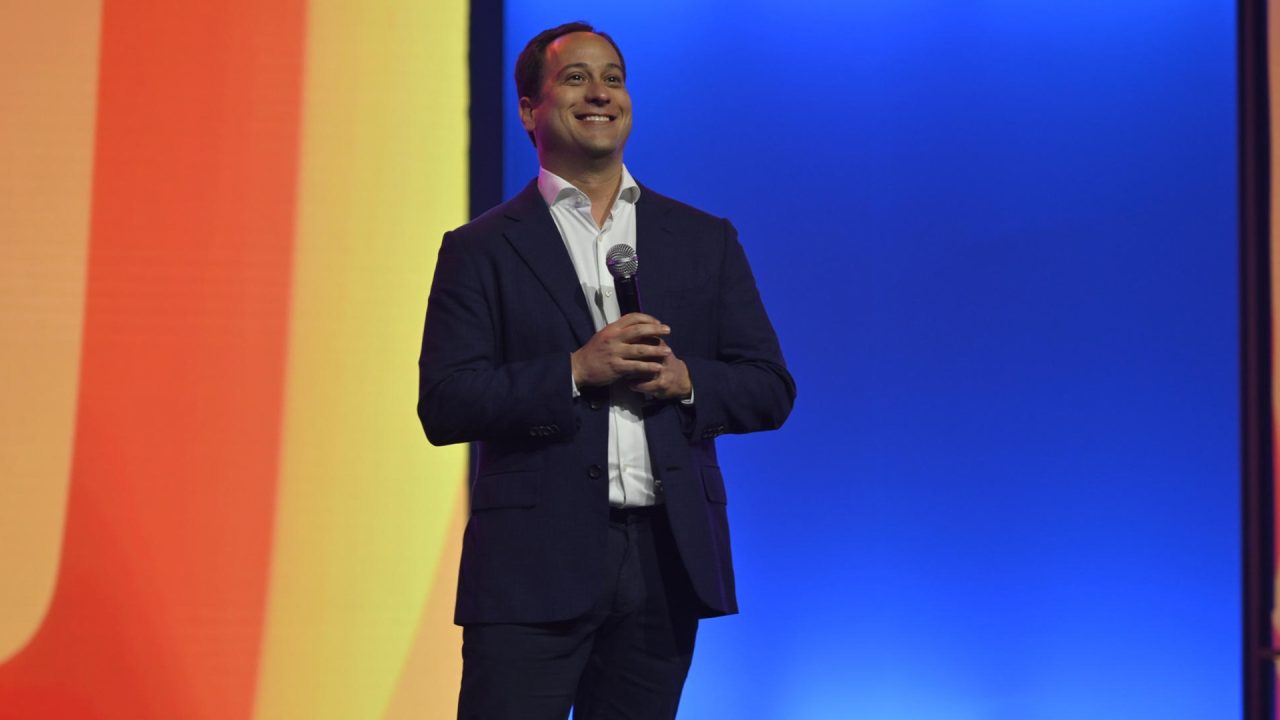 Paulo Marinho, diretor-presidente da Globo, veste terno e segura microfone, sorrindo em palco com fundo colorido em degradê durante evento do conglomerado.