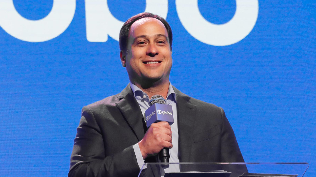 Paulo Marinho sorri enquanto segura microfone da Globo durante apresentação em frente a tela azul com logotipo da empresa
