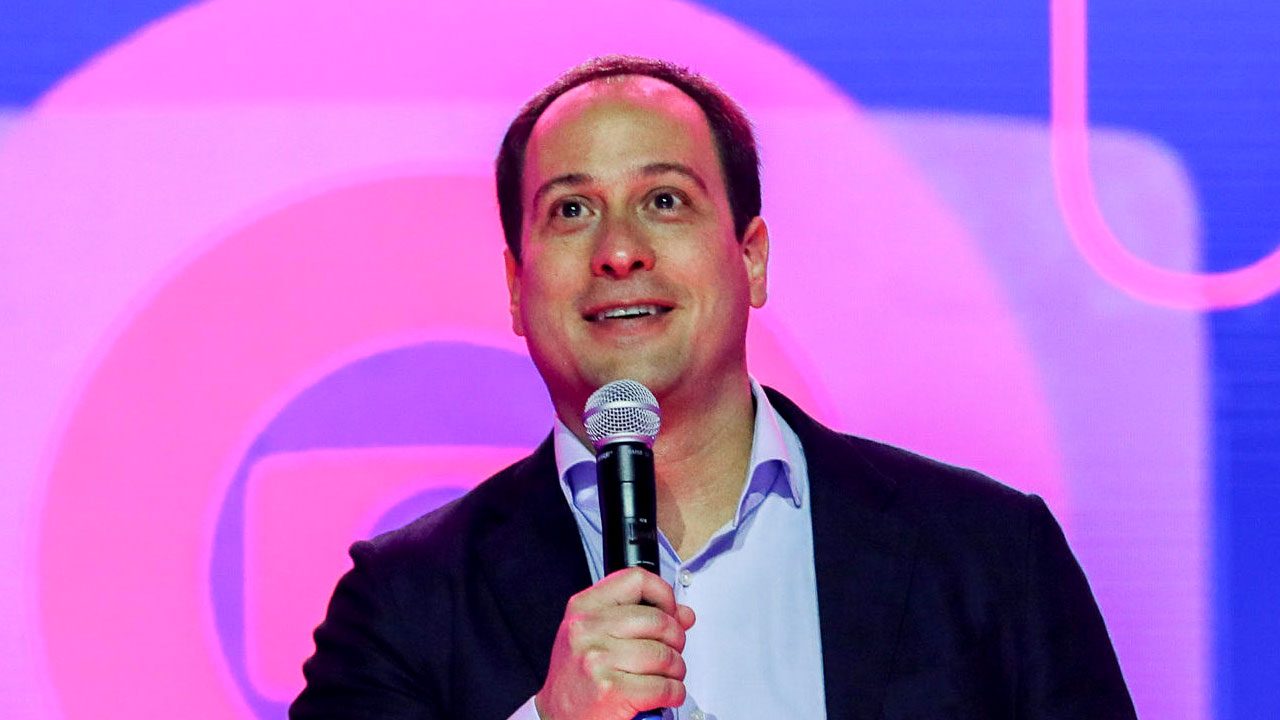 Paulo Marinho fala ao microfone vestindo terno azul marinho e camisa clara em frente a um telão com o logotipo da Globo ao fundo