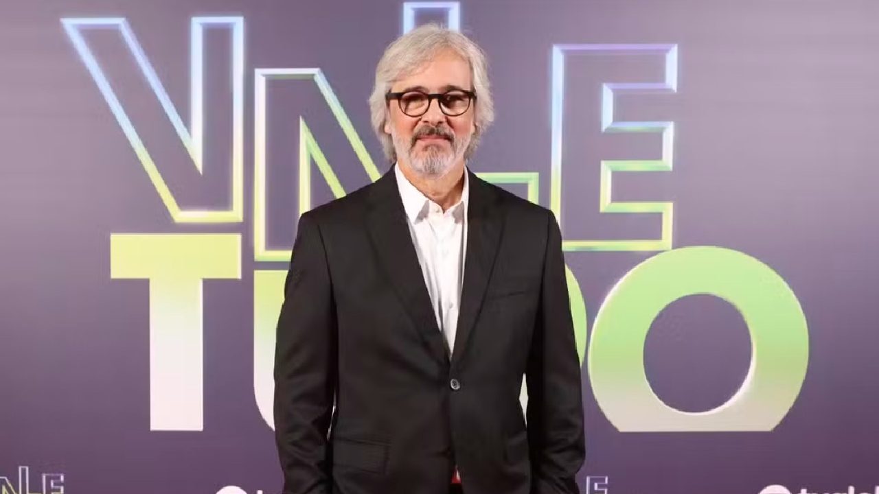 Paulo Silvestrini de terno preto e camisa branca posa diante de painel com logotipo da novela Vale Tudo em evento de lançamento