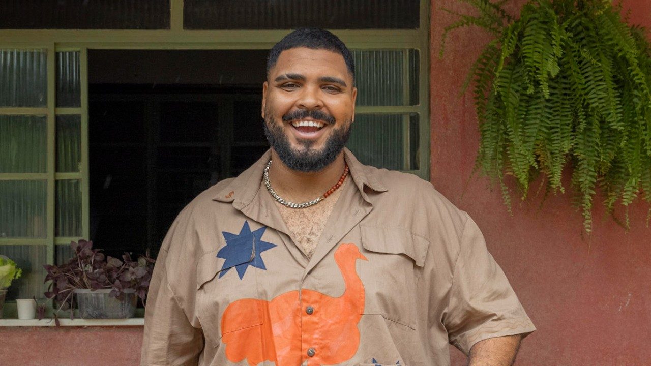 Paulo Vieira sorrindo com camisa estampada diante de janela e parede com samambaia pendurada
