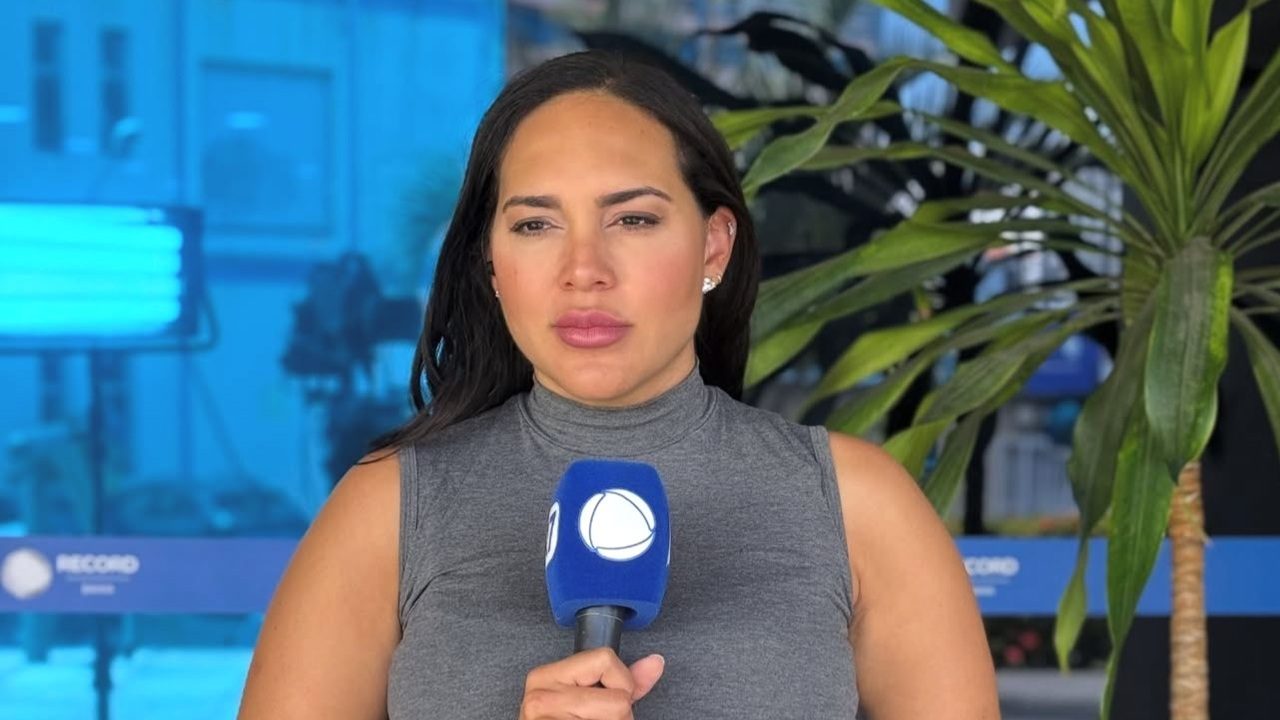 Repórter segura microfone azul com logotipo de emissora, olhando seriamente para a câmera durante gravação em local externo com fundo azul e planta