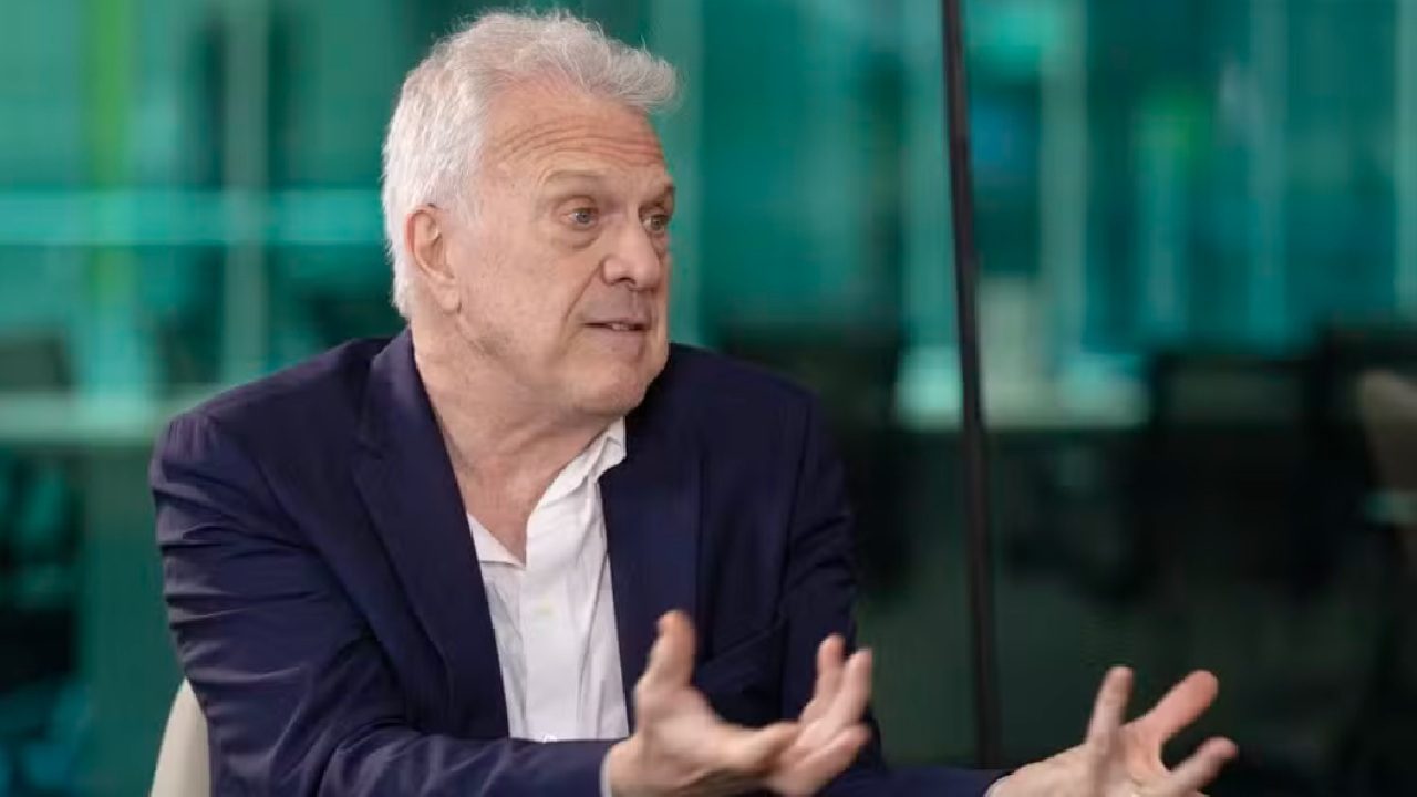 Homem de cabelos grisalhos em terno azul gesticula enquanto conversa em ambiente corporativo com fundo envidraçado