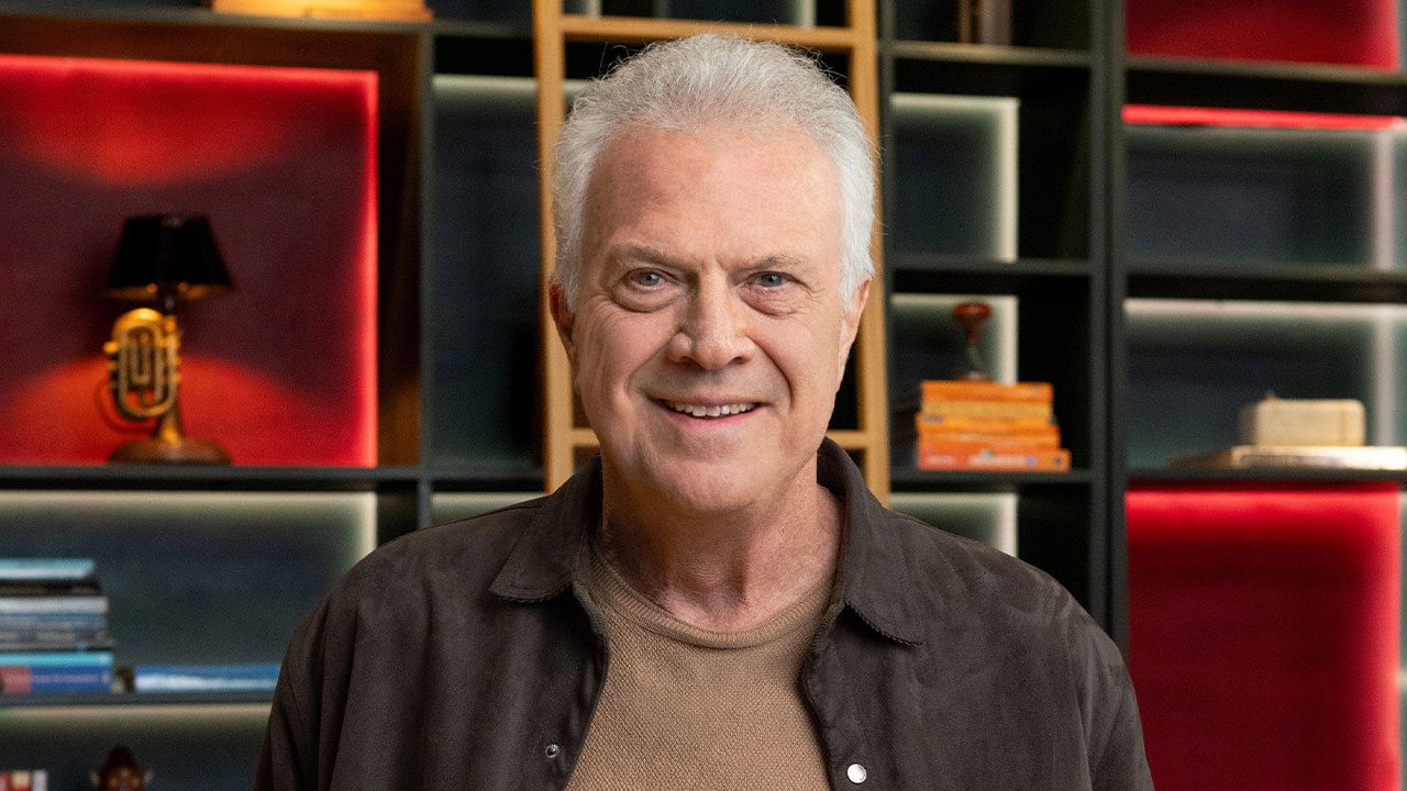 Pedro Bial sorrindo em primeiro plano com cabelo grisalho e jaqueta marrom, diante de uma estante de livros e objetos de decoração