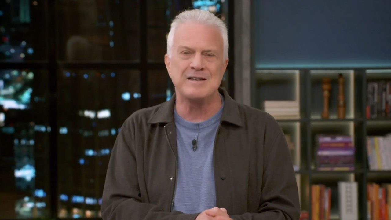 Pedro Bial sorri levemente usando jaqueta marrom e camiseta azul, com as mãos cruzadas e estante de livros ao fundo, em cenário do Conversa com Bial