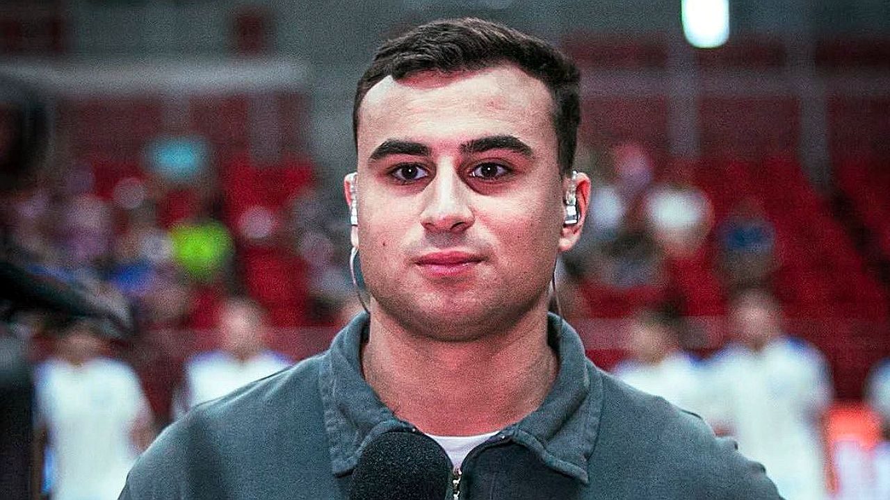 Um homem aparece em primeiro plano durante uma transmissão, olhando diretamente para a câmera. Ele usa um fone de ouvido intra-auricular e segura um microfone, indicando que está trabalhando como repórter ou comentarista. Veste uma blusa de moletom escura. Ao fundo, há uma arquibancada com várias pessoas desfocadas, sugerindo um ambiente esportivo ou ginásio.