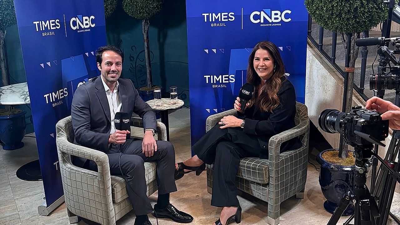 Homem e mulher seguram microfones em entrevista diante de painéis da Times Brasil CNBC