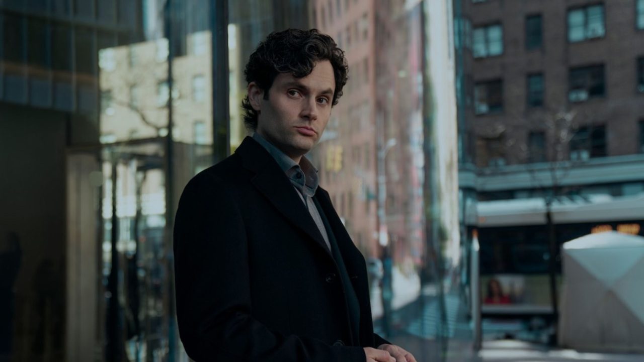 Homem de expressão séria veste casaco escuro e olha fixamente para frente em uma rua urbana, com prédios e reflexos de vidro ao fundo. A iluminação fria e o enquadramento sugerem um clima de mistério ou tensão psicológica