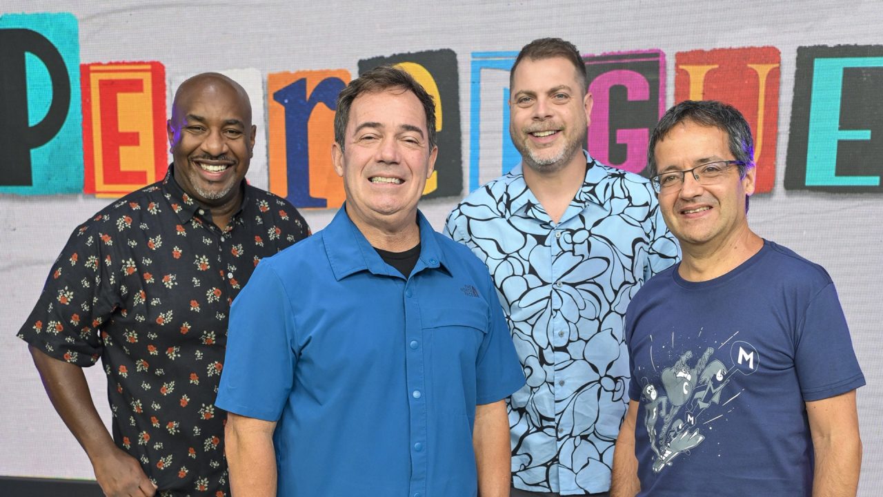 Ângelo Campos, Tatola Godas, Dennys Motta e Ricardo Mendonça posam lado a lado em frente a um painel colorido com a marca do Perrengue na Band. Cada um veste camisa casual diferente — uma estampada escura, uma azul lisa, uma azul com estampa floral e uma camiseta azul com ilustração.