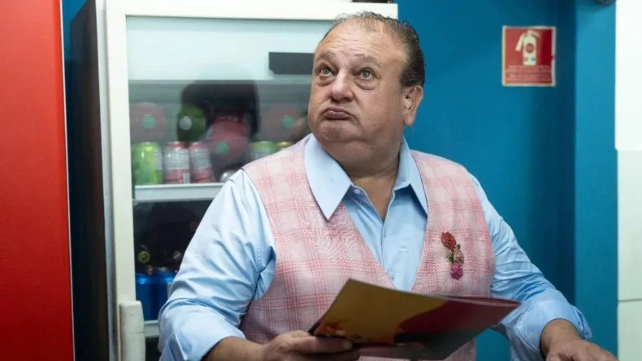 A imagem mostra um homem de colete rosa xadrez e camisa azul-clara, segurando um cardápio ou pasta. Ele está em um ambiente interno, aparentemente uma cozinha ou restaurante, com uma geladeira ao fundo contendo bebidas. Sua expressão parece pensativa ou curiosa, com o olhar voltado para cima