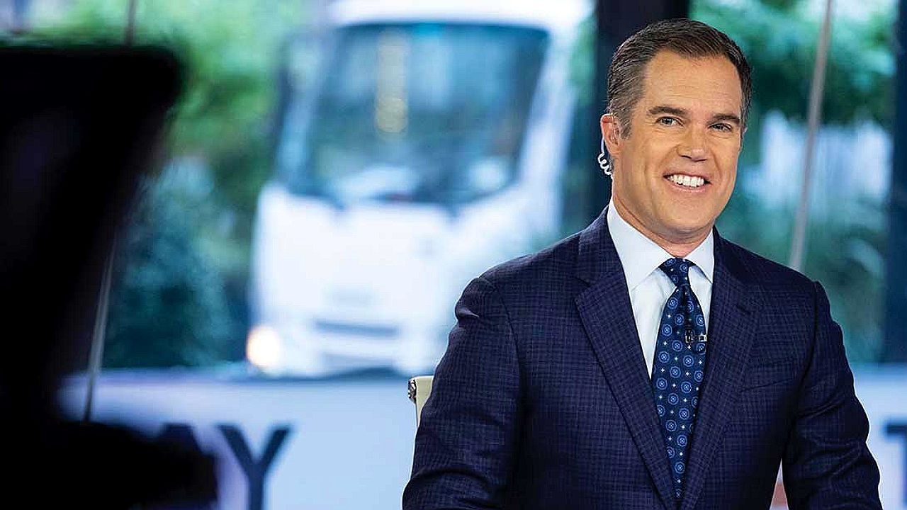 Homem de terno e gravata sorri durante apresentação em estúdio de telejornal com telão ao fundo