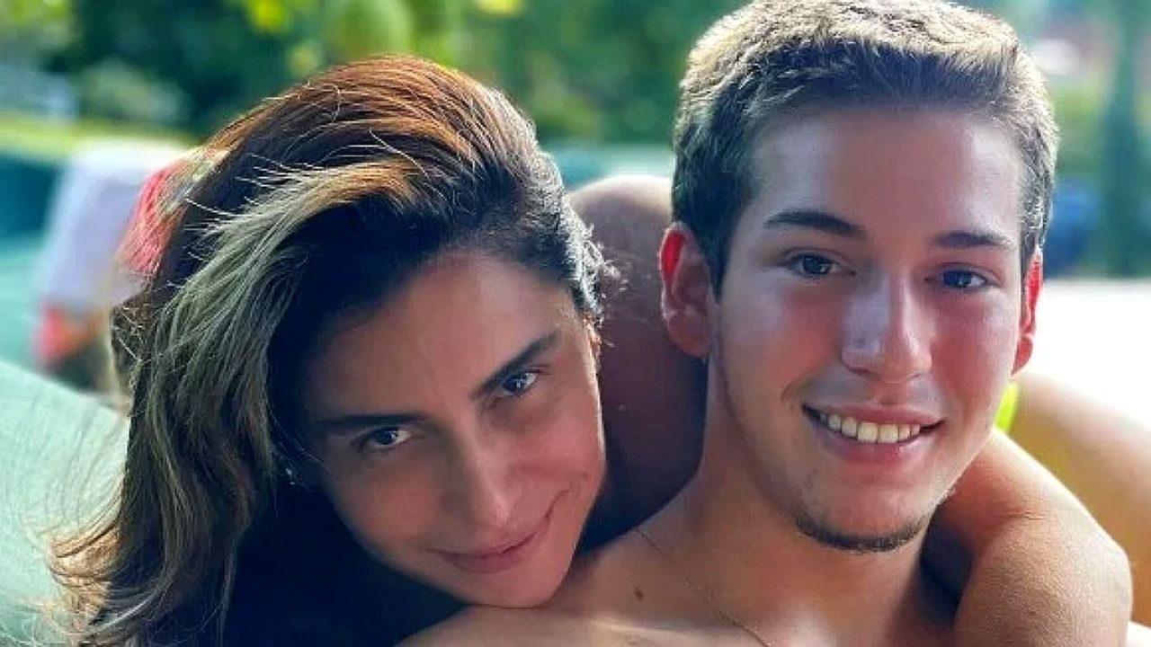 Na imagem aparecem duas pessoas: um homem e uma mulher. Eles estão abraçados, com a mulher apoiada nas costas do homem, ambos sorrindo para a câmera. Parece uma foto casual ao ar livre, com fundo desfocado de vegetação e carros.