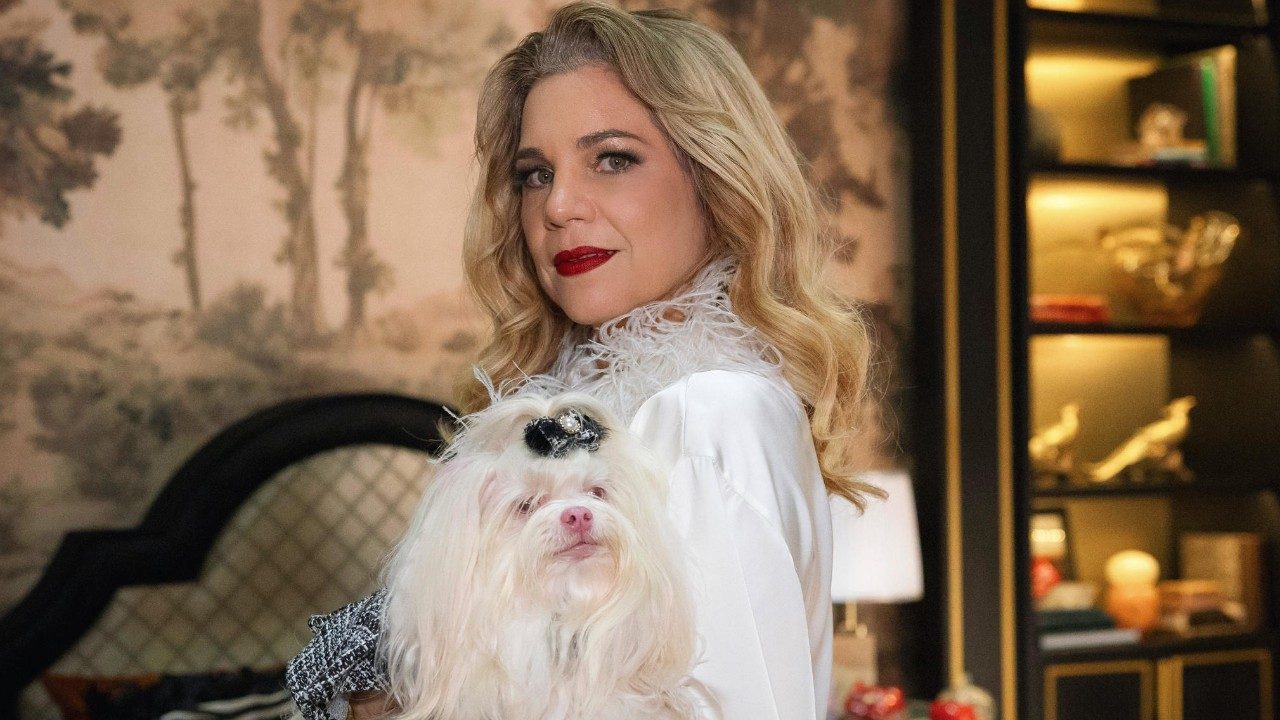 Pilar (Isabel Teixeira) segura cachorro branco no colo, veste roupa branca com gola de plumas em cena de Quem Ama Cuida