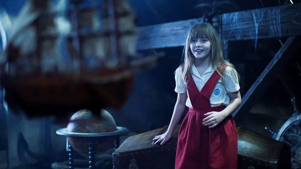 Menina com uniforme escolar vermelho e branco sorri em cenário de fantasia com navio e objetos antigos sob iluminação azulada