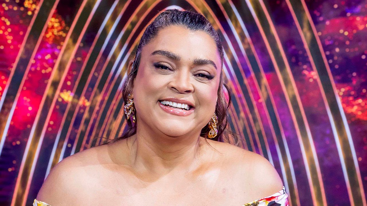 Preta Gil sorrindo como jurada no palco do The Masked Singer Brasil com fundo colorido iluminado