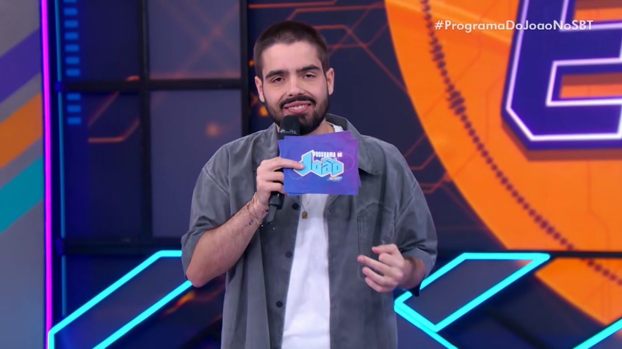 João Silva apresenta o Programa do João no SBT com microfone e card do programa em um estúdio colorido.