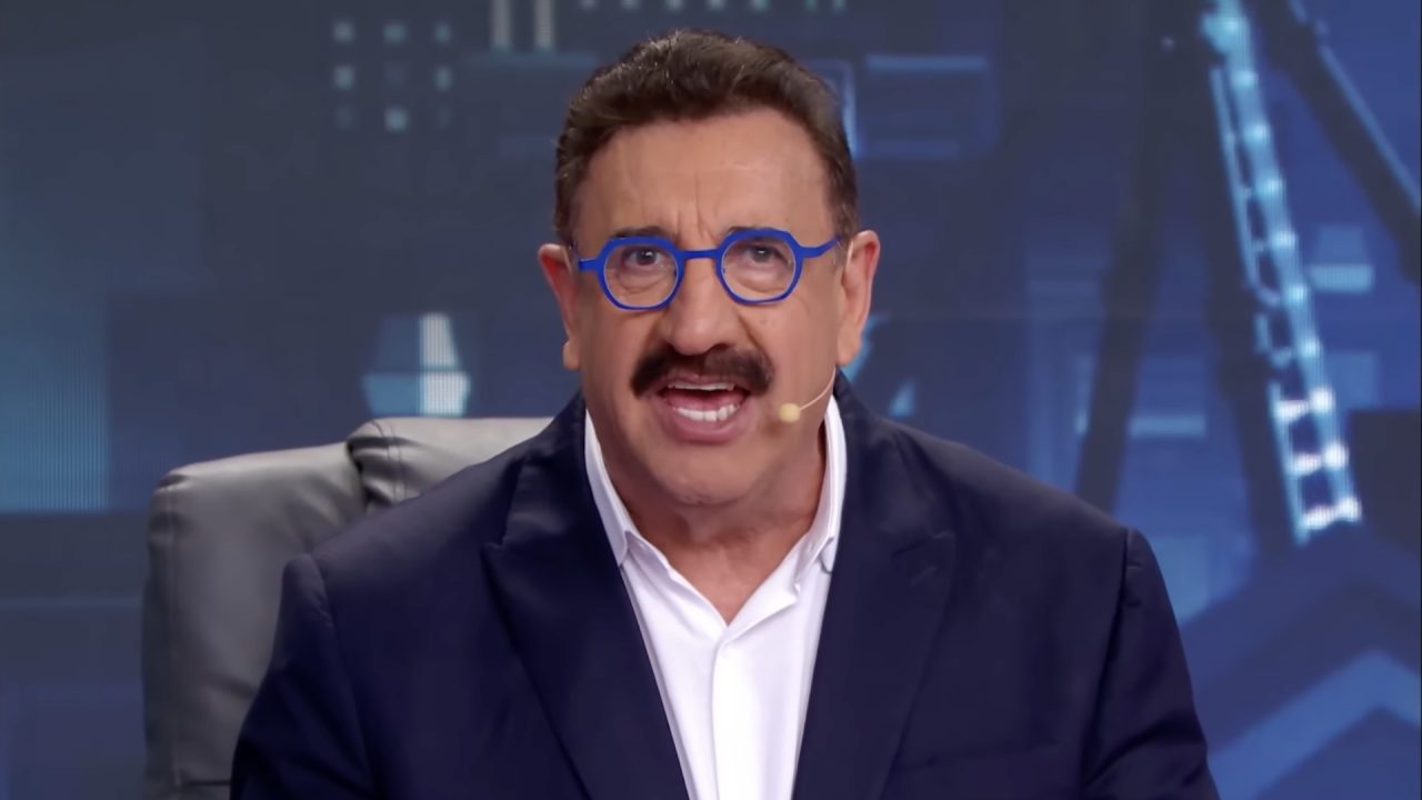 Ratinho, usando terno preto e um óculos de armação azul, apresenta seu programa no SBT, com um cenário composto por telões de LED com imagens de cidades.