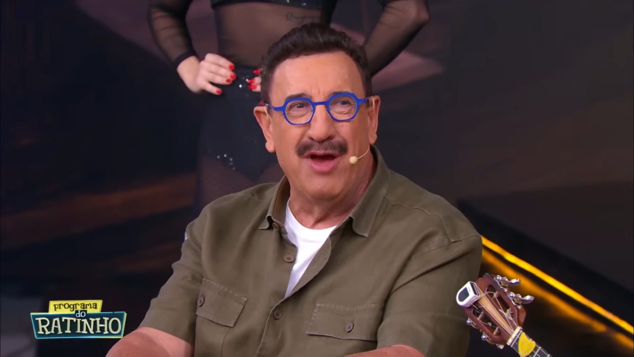 Ratinho fala durante o Programa do Ratinho no SBT com microfone e músico ao fundo em estúdio.