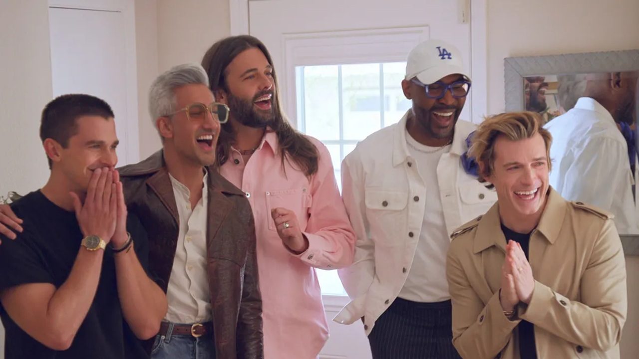 Cinco homens estão lado a lado em um ambiente interno iluminado, sorrindo e rindo com expressões de empolgação e alegria. Um deles usa camisa preta e relógio dourado, outro veste jaqueta marrom com óculos grandes, o terceiro usa camisa rosa, o quarto veste jaqueta branca e boné branco, e o quinto usa sobretudo bege. Ao fundo há uma porta branca com vidro e um espelho refletindo parte da cena.