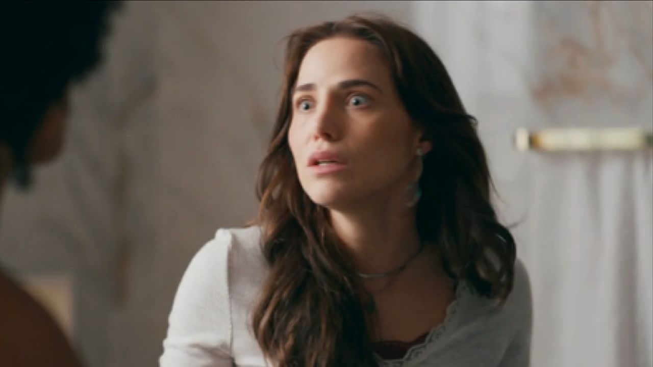 Mulher de cabelos escuros arregala os olhos com expressão de choque durante conversa tensa em cena de novela ambientada em residência elegante