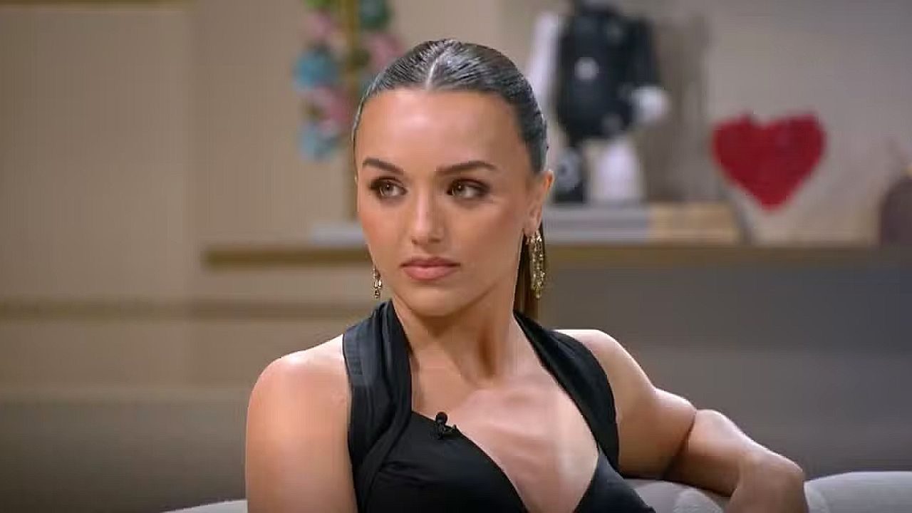 Mulher de vestido preto com cabelo preso participa de entrevista em estúdio com expressão séria em programa de TV