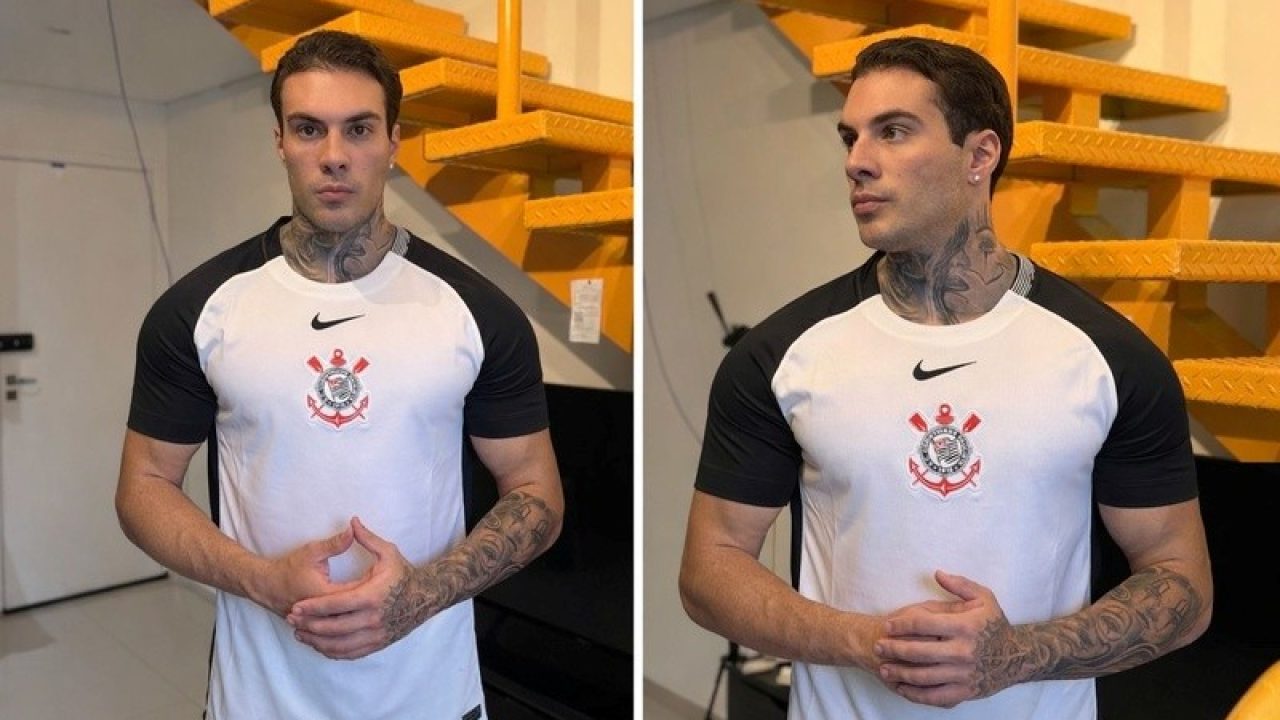 Rafa Marttinz veste camisa branca do Corinthians com mangas pretas, exibe tatuagens no pescoço e braços, posicionado à frente de uma escada amarela clara