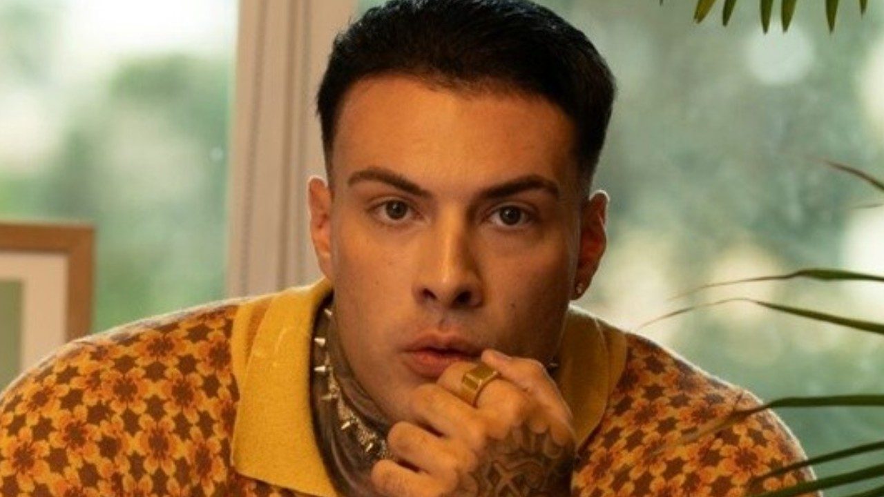 Rafa Marttinz olha surpreso com a mão na boca em ambiente interno com janela e plantas ao fundo