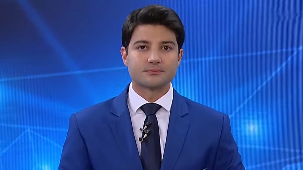 Rafael Algarte veste terno azul e gravata escura enquanto apresenta telejornal da Record News em estúdio com fundo azul geométrico