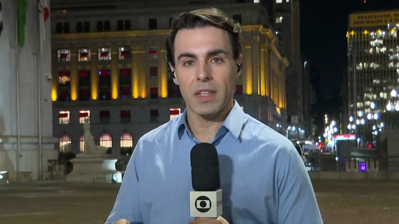 Rafael Colombo com microfone da Globo em frente a prédio iluminado durante link ao vivo no centro de São Paulo à noite
