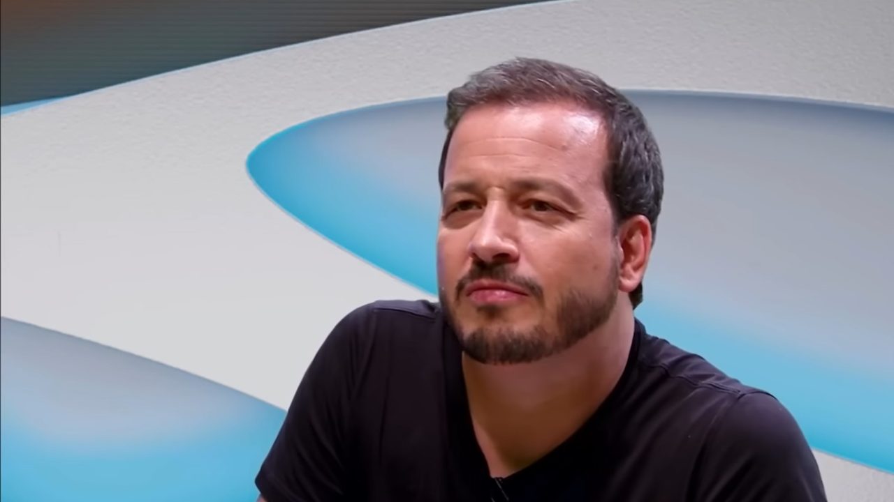 A imagem mostra o humorista e apresentador Rafael Cortez em um cenário moderno, com fundo abstrato em tons de azul e branco. Ele veste uma camiseta preta e tem uma expressão séria e reflexiva, olhando para algo fora do quadro. Seu cabelo está curto e levemente ondulado, acompanhado de uma barba bem cuidada