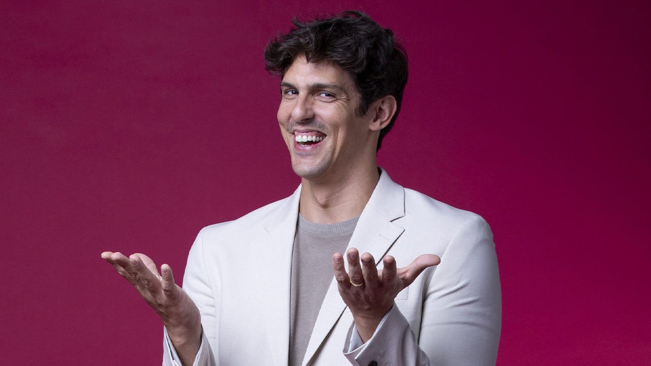 Rafael Infante sorri e gesticula com as mãos enquanto posa para foto, vestindo blazer claro e camisa bege, diante de um fundo roxo