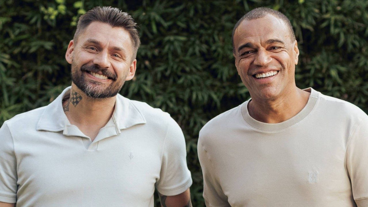 Rafael Sóbis e Denilson sorriem lado a lado ao ar livre com fundo de vegetação em foto posada