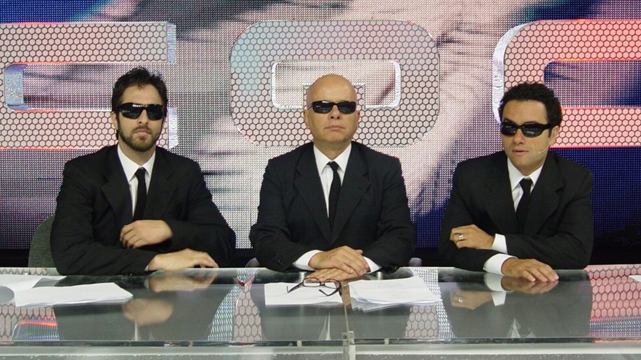 Rafinha Bastos, Marcelo Tas e Marco Luque com terno preto e óculos escuros na bancada do CQC, programa da Band, em sua formação original