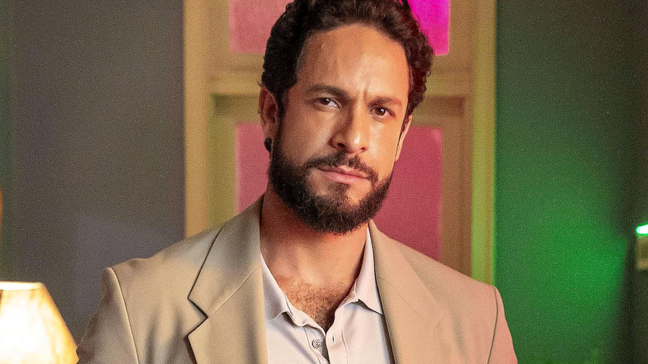 Ator de barba e blazer bege com expressão séria em cena de novela ambientada em casa noturna