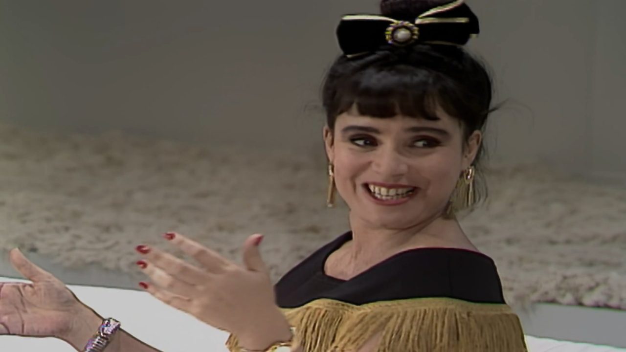 A imagem mostra uma personagem de Rainha da Sucata sorridente, com penteado preso adornado por um laço preto e dourado, brincos longos e vestido com franjas douradas. Ela parece gesticulando com entusiasmo, sugerindo uma cena leve e descontraída