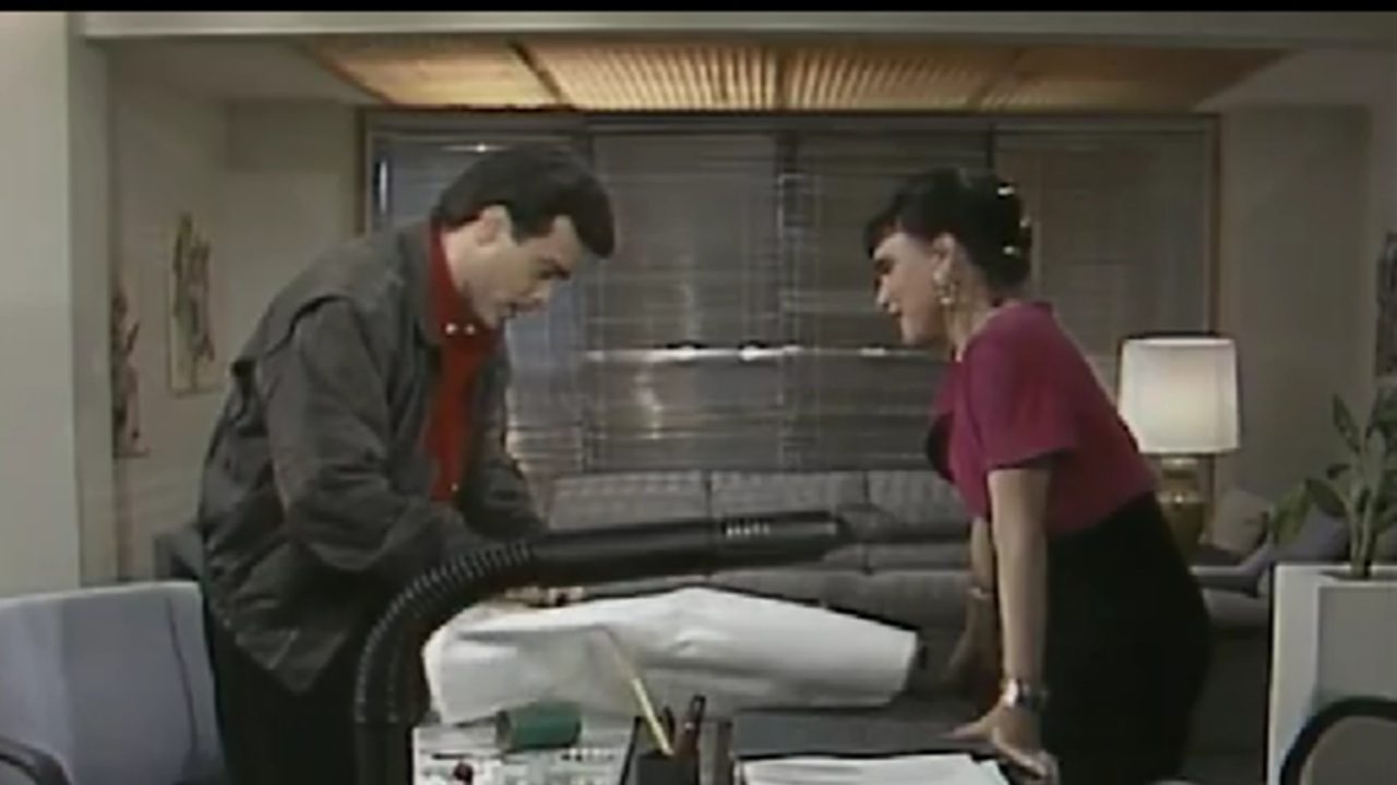 Homem e mulher conversam em um escritório elegante, inclinados sobre uma mesa repleta de papéis e materiais de trabalho. O homem segura um grande documento ou planta dobrada, enquanto a mulher observa atentamente. Ao fundo, há um sofá, luminária e persianas fechadas, compondo um ambiente corporativo dos anos 1980 ou 1990.