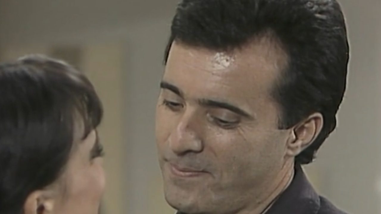 Close em um homem olhando de forma suave para uma mulher que aparece parcialmente de costas. Ambos estão em um ambiente interno, e a cena transmite um momento de intimidade e conversa próxima.