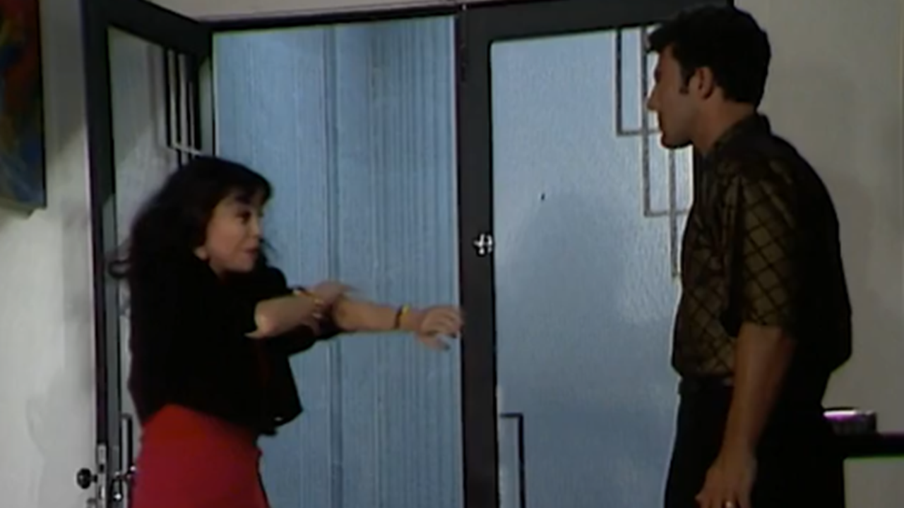 Mulher faz gesto enfático com os braços diante de um homem, em ambiente interno iluminado, próximo a uma porta de vidro; a cena sugere uma conversa intensa ou discussão