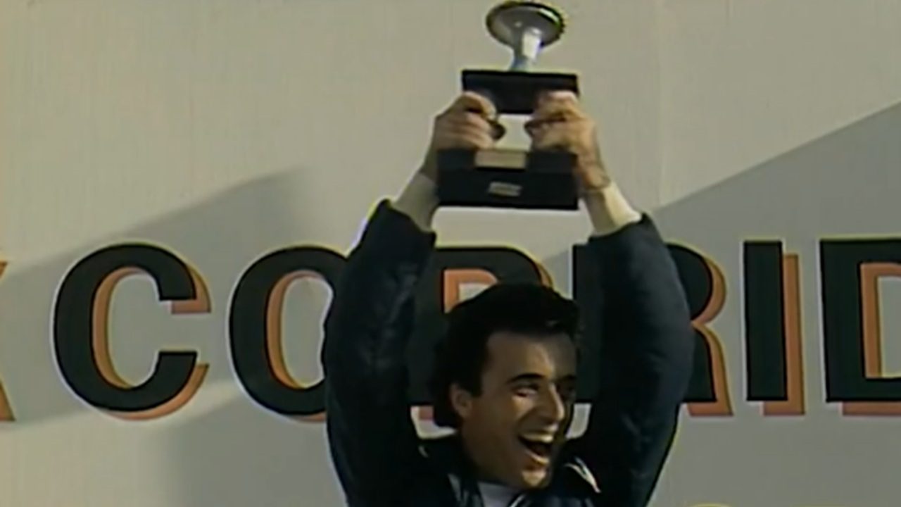 A imagem mostra um homem sorridente erguendo um troféu acima da cabeça em um pódio de premiação. Ele veste um macacão de corrida azul e parece comemorar uma vitória esportiva. Ao fundo, há parte de um painel com letras grandes que indicam um evento competitivo